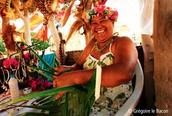 Femme polynésienne souriante, couronne de fleurs sur la tête, tressant des feuilles de palmier.