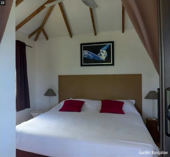 Chambre d'un Garden Bungalow du Havaiki Lodge avec lit double blanc, coussins rouges et charpente en bois.