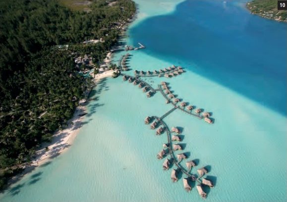 Vue aérienne du Bora Bora by Pearl Resorts avec ses bungalows sur pilotis dans le lagon turquoise et le motu boisé.