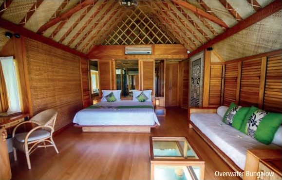 Intérieur d’un bungalow sur pilotis en bois avec lit double, canapé à coussins verts et table basse vitrée.