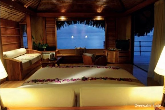 Intérieur d'une suite sur pilotis au crépuscule, lit décoré de fleurs face à une baie vitrée ouverte sur le lagon.