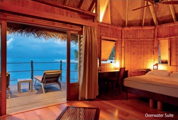 Intérieur boisé d'une Overwater Suite avec lit, bureau et terrasse ouverte sur le lagon.