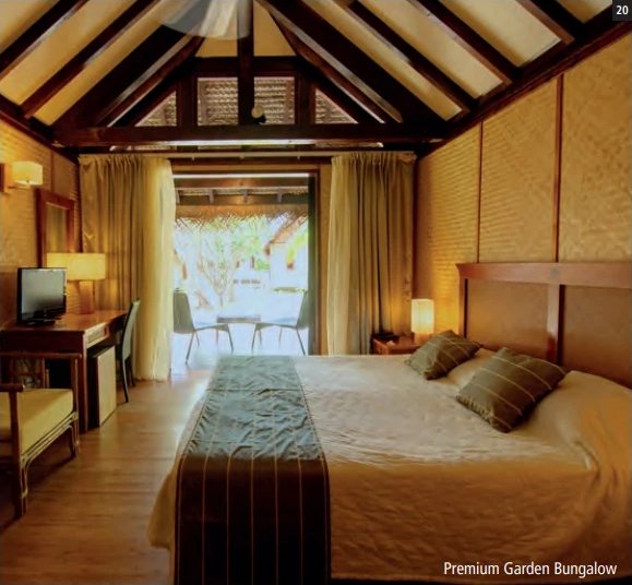 Chambre d'un bungalow avec grand lit, charpente apparente, rideaux dorés et porte-fenêtre ouverte sur la terrasse.