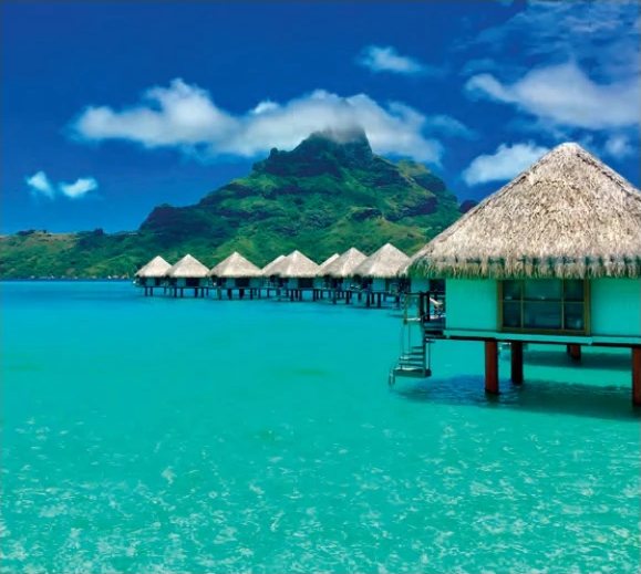 Bungalows sur pilotis au toit de chaume sur un lagon turquoise, avec une montagne verdoyante en arrière-plan.