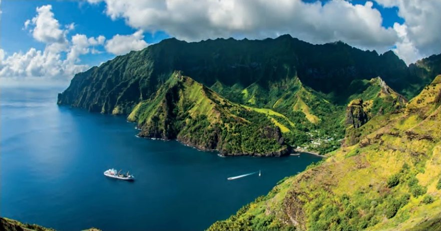 Baie profonde bordée de montagnes verdoyantes et escarpées, avec un navire et un bateau rapide sur la mer bleue.