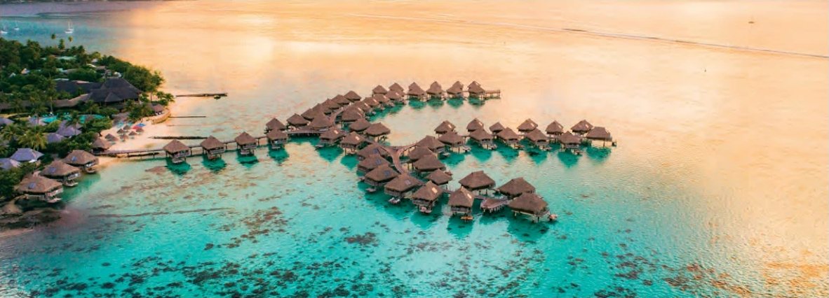 Vue aérienne d'un hôtel avec bungalows sur pilotis dans un lagon turquoise au coucher du soleil.