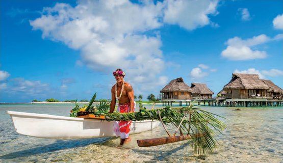 Un Polynésien couronné de fleurs pousse une pirogue à balancier décorée de palmes dans le lagon, devant des bungalows sur pilotis.