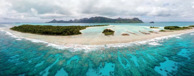 Vue aérienne d’un motu bordé de sable blanc et d’un récif corallien turquoise, avec une île montagneuse à l’horizon.