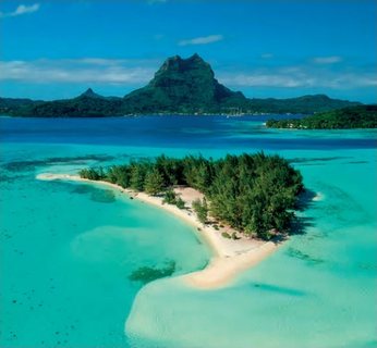 Vue aérienne d'un motu boisé bordé de sable blanc dans un lagon turquoise, avec la silhouette montagneuse de Bora Bora en arrière-plan.