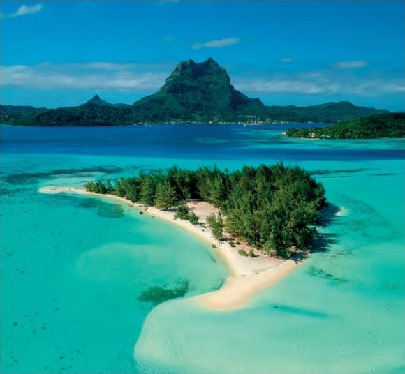 Vue aérienne d'un motu boisé bordé de sable blanc dans un lagon turquoise, avec la silhouette montagneuse de Bora Bora en arrière-plan.